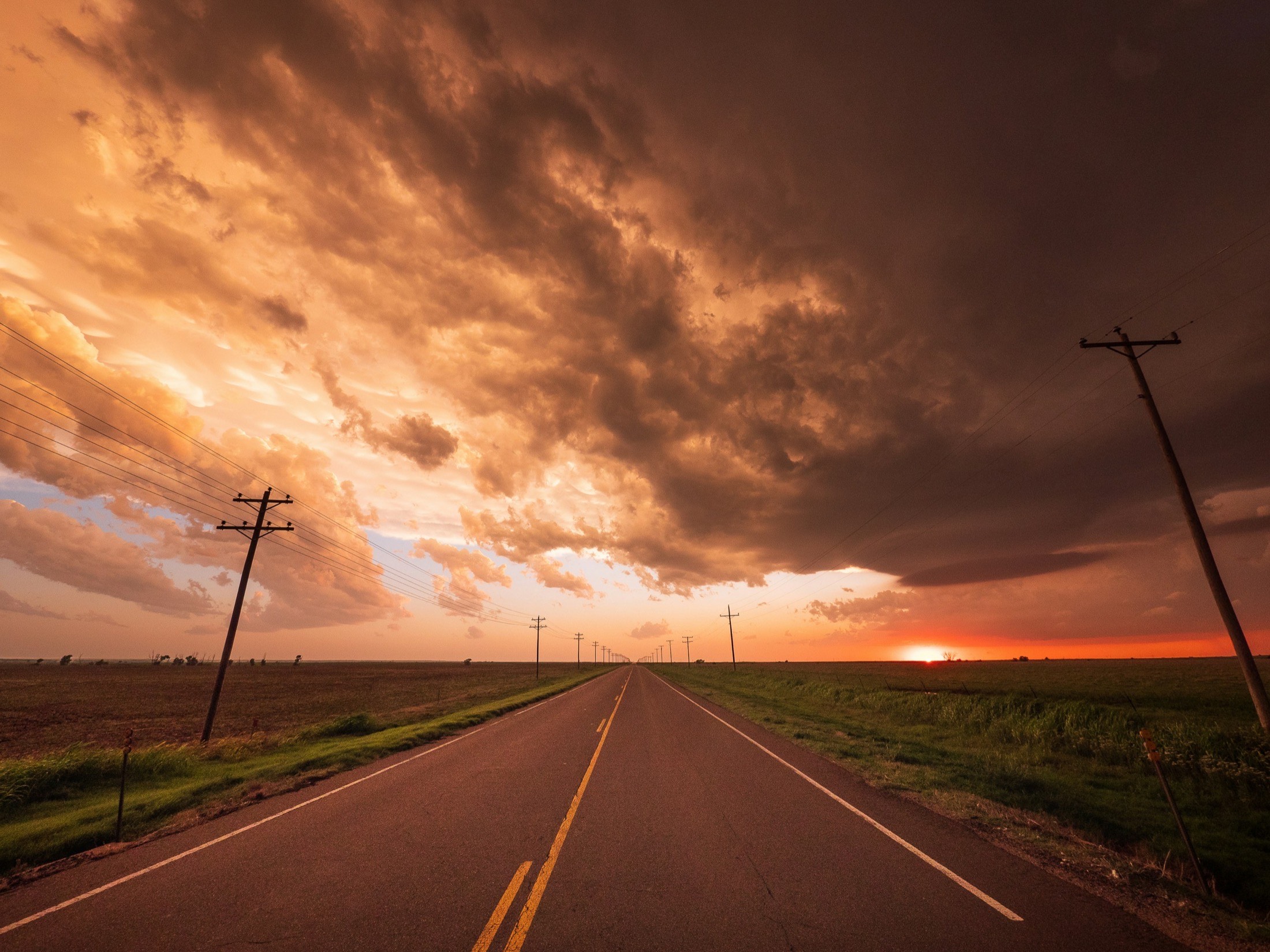 An open two-lane highway stretching through flat Oklahoma farmland under a dramatic sunset sky.